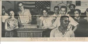 Nine young white people stand behind a black man who is singing or speaking. A backwards United States flag hangs on the wall in the background. 