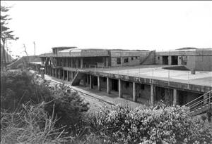 A long concrete structure with small windows along the top and steel doors along the ground floor. Shrubs on a hillside in the foreground. 
