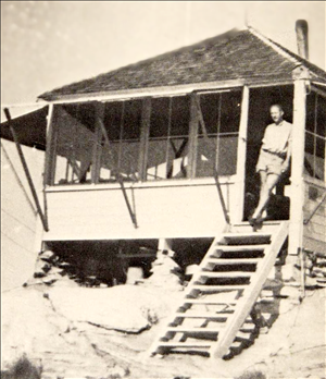 A person stands in the doorway looking out from a one room wooden structure with a peaked roof. The window shutters are propped open and there are many windows. Wooden stairs lead down from the open door. 