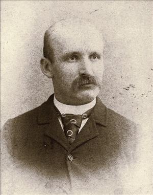 Photographic portrait of a middle aged man who is bald with a mustache and is wearing a dark suit, white shirt, and patterned tie.
