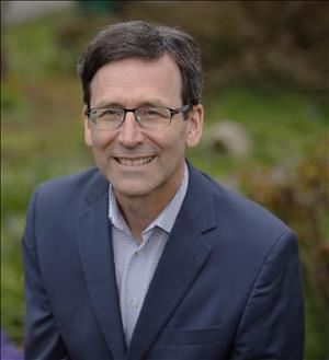 Head and shoulders portrait of a man with glasses in sports jacket and open-collared shirt, out-of-focus outdoor setting in background 