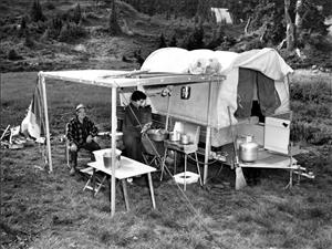 Two people next to their camper in a meadow at the base of a forested slope. The camper is a trailer with a round canvas top. A four legged shelter next to the camper covers a camp stove and table. The two campers are beneath the shelter. One is seated and the other is cooking. 