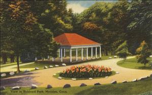 A postcard of a picnic structure in a park. The structure has a red roof help up by white beams and is surrounded by trees. In the foreground, a traffic circle full of orange flowers in the middle of a dirt road. 