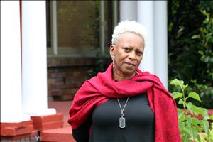 A head and shoulders portrait of a Black woman in a red scarf. She is standing in front of white columns. 