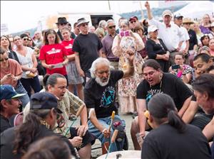 Indigenous drummers seated in a circle outdoors with a large crowd assembled behind them