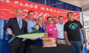 Seven adults of different genders and races pose for the camera holding a swordfish prop. There is a cake on the table in front of them and a red banner hanging behind them