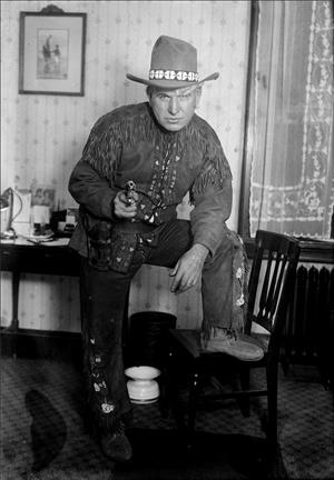 A man in cowboy attire and a large cowboy hat stands with one foot up on a chair, an arm across his knee. He is pointing a pistol at the camera. He is in a small room with lace curtains and a spittoon or chamber pot on the floor. 