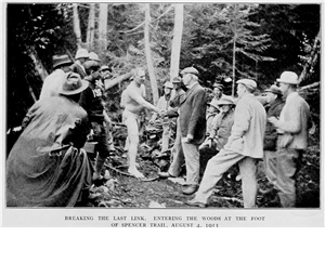 A man wearing only underpants shakes the hand of a man who is fully dressed. They are in the forest and are surrounded by several other people all looking at the man in underpants. 