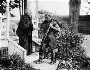 A bearded man wearing animal skins and carrying sticks shakes the hand of an elderly woman who is standing on stairs to a porch.