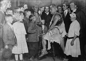 A white man wearing animal skins sits on a stool in front of an indigenous man with long hair wearing a suit jacket. They are surrounded by children leaning in towards them and smiling. 