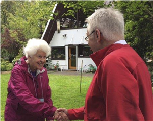 A smiling elderly white woman shakes someone's hand in front of an A frame house surrounded by trees. 