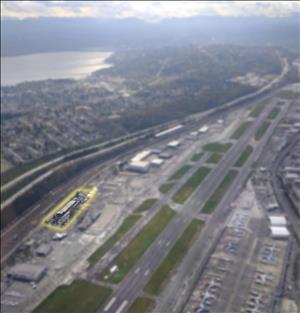 Birds eye view of a runway with parked airplanes to the right. To the left is a freeway and rolling hills of residential neighborhoods. The image is blurry except for one building next to the runway with a yellow line around it. 