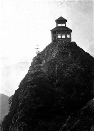 A small one room wooden structure on top of a steep peak. The structure has windows on all sides and a peaked cupola roof that also has windows on all sides. A person is standing in the open doorway. 