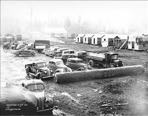 Cars and trucks parked in the mud in front of small wooden cabins and tents. 