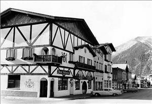 A large three story hotel with a white facade, Tudor style decorations, and wooden balconies. A line of decorative buildings are next to the hotel, with large snowy mountains in the background, and cars parked in front. 