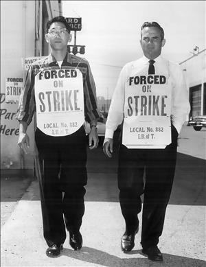 Two men, one asian one white, walking with signs around their neck that say forced on strike. Industrial buildings are behind them. 