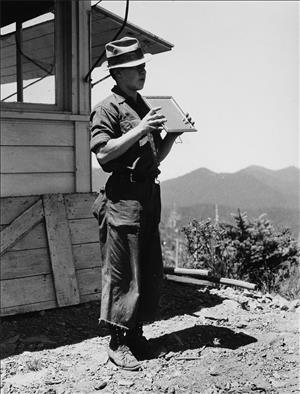 A person in a floppy hat stands outside a wooden structure holding a mirror in front of them facing away from them. In the background are rolling hills. 