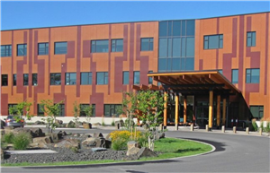 Color photo of front of modern 3-story office building, orange with brick-red designs and blue glass windows, roofed entrance at right, and driveway with circular park in front 
