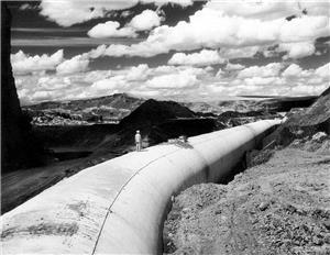 A man in a white suit stands on a long white pipe that snakes through mountains under a cloudy sky