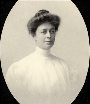 A portrait of a white woman with long hair up in a loose bun and wearing a white blouse with a high collar covering her neck. 