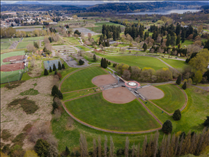 Bird's eye view of a large park with forested hillsides and a lake in the distant background. Circular sports fields are surrounded by mowed grass fields and paths. 
