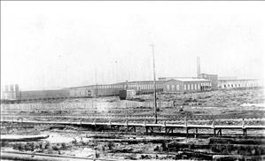 Wooden factory buildings in rows. Train tracks in the foreground. 