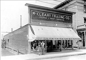 A one story storefront with a tall front display and awning that says McCleary Trading Co. A man with a broom stands on the sidewalk in front of the store. 