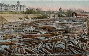Antique postcard tinted in pastel colors shows a pond full of floating logs and a large white institutional building on the banks of the pond. 