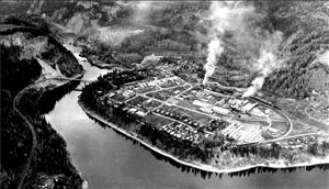A birds eye view of a small town and mill on the shore of a large river. Smoke rises from smokestacks. 