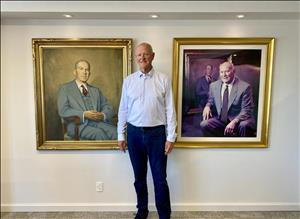 A tall white man stands between two portraits. On the left is a painting of a man in a light colored suit and red tie, leaning back in an armchair. On the right is a photographic portrait of a man in a suit seated in front of the painting that is on the left. 