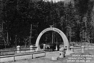 A stone archway over a road through the forest. On the archway it says Moran State Park. A car is parked under the arch with passengers standing on either side of it. 