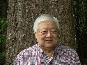 An elderly Asian man stands in front of a tree. 