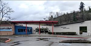 A one story commercial building with slanted roofs and a red entryway. A forested hillside in the background. 