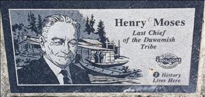 A stone plaque. Carved into the plaque is the face of an elderly indigenous man in a suit and tie with a canoe, cabin, and forested hill behind him. The words on the plaque say henry moses last chief of the duwamish tribe history lives here. 