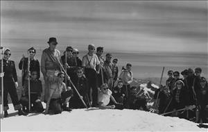 A large group of people in sunglasses holding walking sticks posing together on a snowy slope with nothing but sky behind them. 