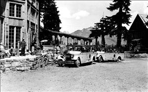 Two nineteen twenties era automobiles with several rows of seats and open sides. They are parked in front of a building with tall windows. They are both full of people. Mountain peaks and forest in the background. 