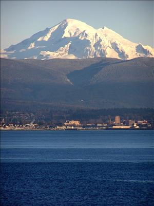 A snow capped mountain peak towers over a coastal city and its waterfront. Rolling tree covered hills are between the mountain and the city. 