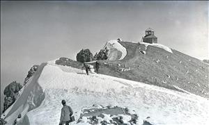 Three hikers on a snowy ridge. Above them is a small wooden structure with a peaked cupola on the roof.
