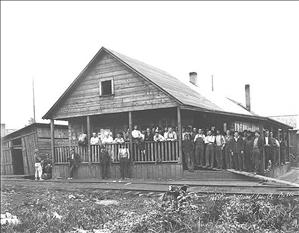 A large group of men in work clothes stand on the porch of a simple wooden building next to railroad tracks. 