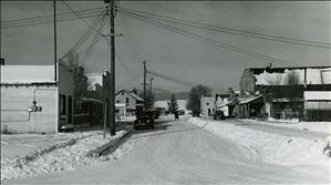 Black and white photo looking down snow-covered street with a few parked cars and storefronts on both sides