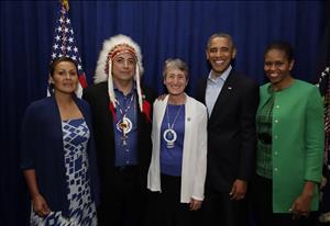 Five people stand close together posing for the camera in front of a blue backdrop. One of them wears traditional native headdress. 