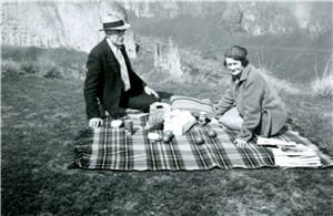 A man and woman sit on a plaid picnic blanket with fruits and other food items spread out on the blanket. Behind them are rocky bluffs. 