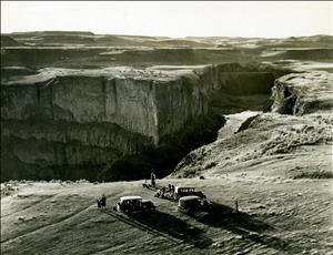 A bird's eye view of a deep canyon with nineteen twenties era automobiles parked on the canyon's edge.  A few people are engaged in various tasks next to the cars. In the distance, rolling plains stretch to the horizon. 