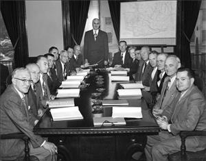 A long table with men in suits on both sides. They are all looking at the camera. One man stands at the head of the table. One woman peeks from behind all the men to look at the camera too. There are books and ashtrays on the table and a map on an easel beside the table.