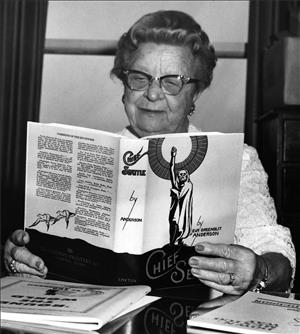 A woman in glasses reads a book that is propped up on a desk. the book cover says Chief Seattle and has an artwork of a man wrapped in a blanket holding his hand above his head. 