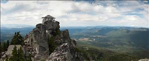 A one room structure with windows on all sides perched on top of a steep rocky peak below which green hills stretch to the horizon. 