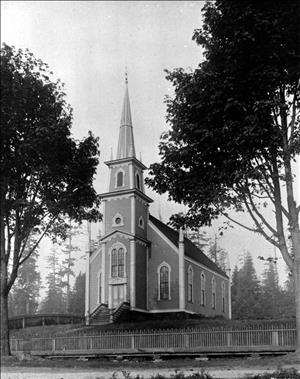 A small church in a clearing in the woods. The church has a tall steeple and tall arched windows. A wooden picket fence wraps around the church. There are trees all around. 