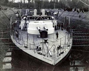 A large white boat in a small dock, surrounded by people, and a small brick building with a smoke stack. In the front are smaller boats, and calm water. 
