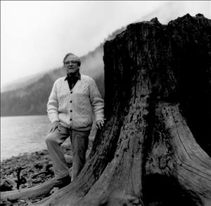 A man stands next to a large tree stump at the water's edge. Fog drifts over a forested hillside behind him. 