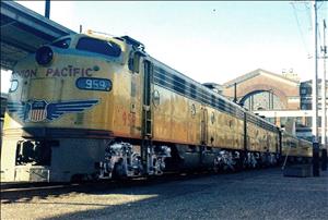 A yellow train locomotive sits on the tracks in front of a large brick building with a peaked roof. 